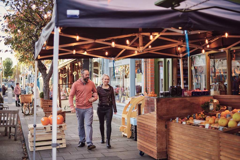 A couple stroll past a street market with fruit stands and autumn decorations in lively Glasthule village.