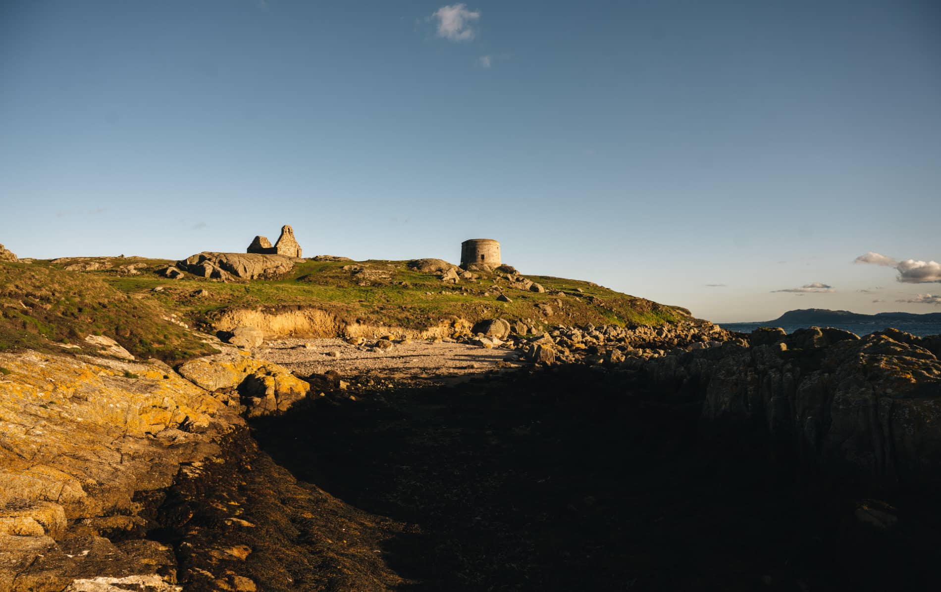 Martello tower and ruins on grassy Dalkey Island viewed from rocky shore with sea in foreground.