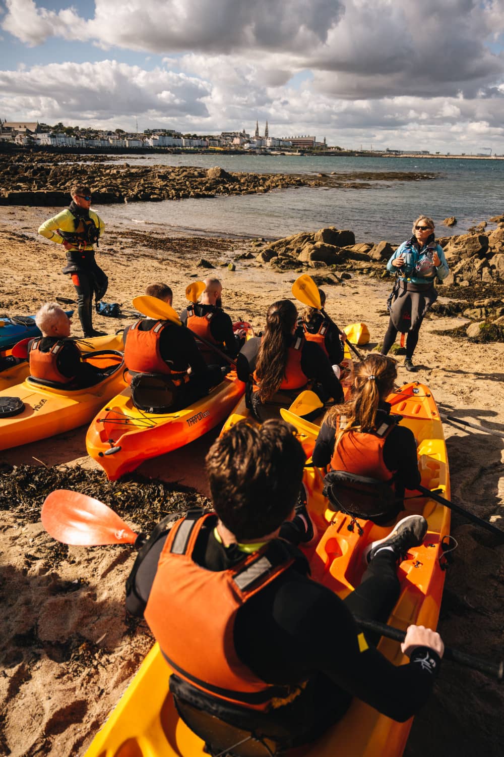 Instructor explaining kayaking basics to a group wearing life jackets on the shore near Dalkey Island.