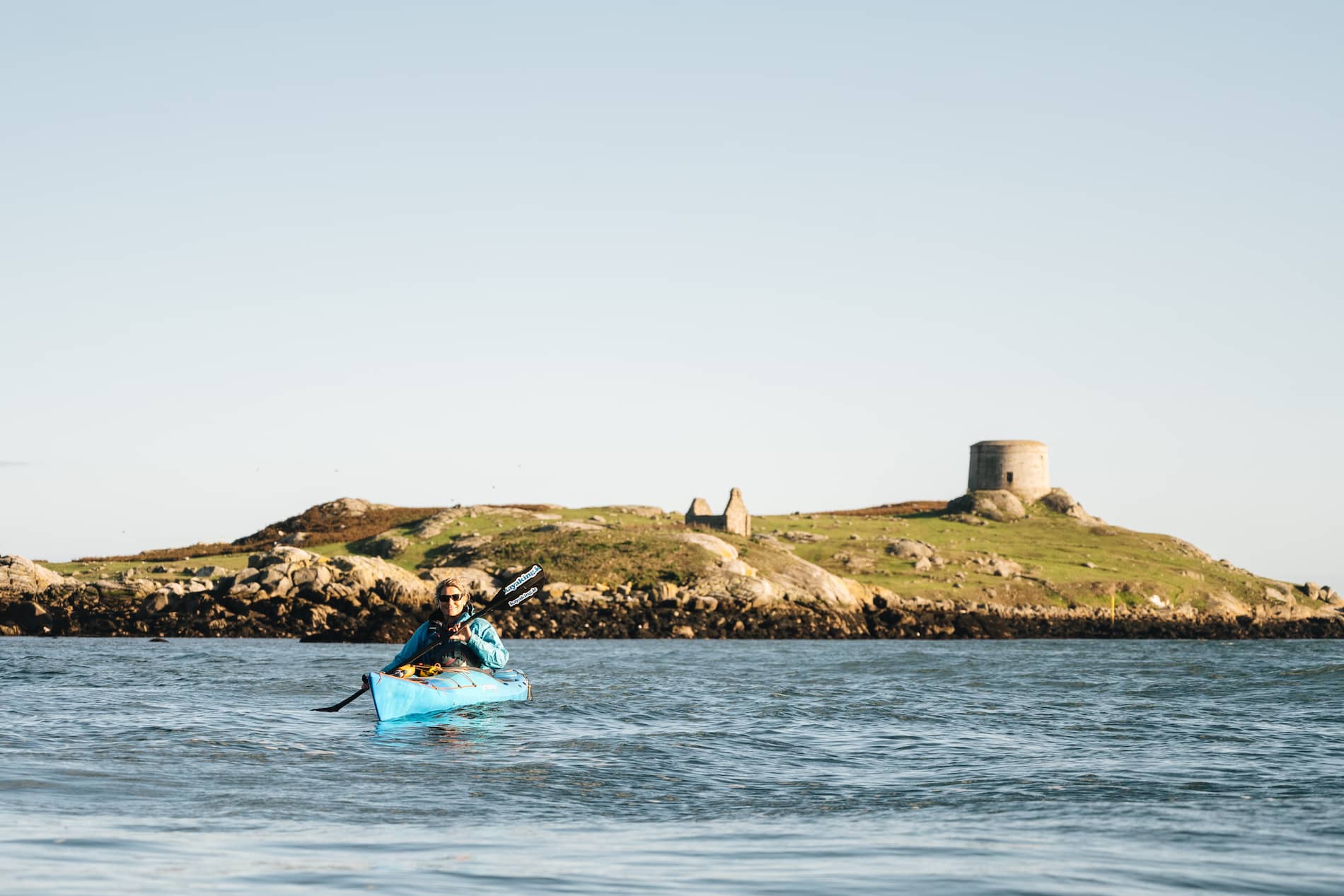 Person kayaking in blue kayak near Dalkey Island Martello tower during clear weather.