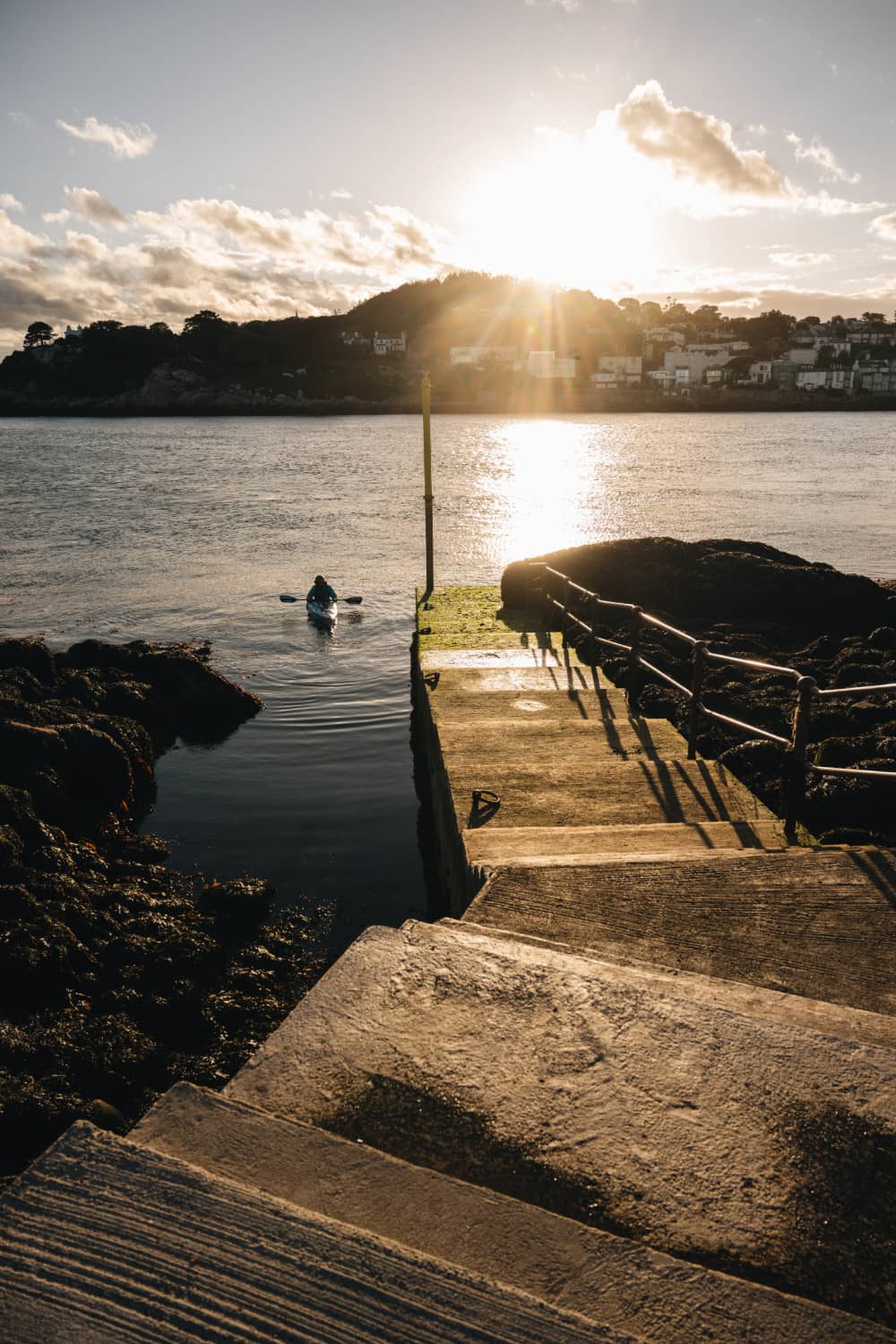Kayaker paddling toward mainland Dublin from Dalkey Island jetty under evening sun.