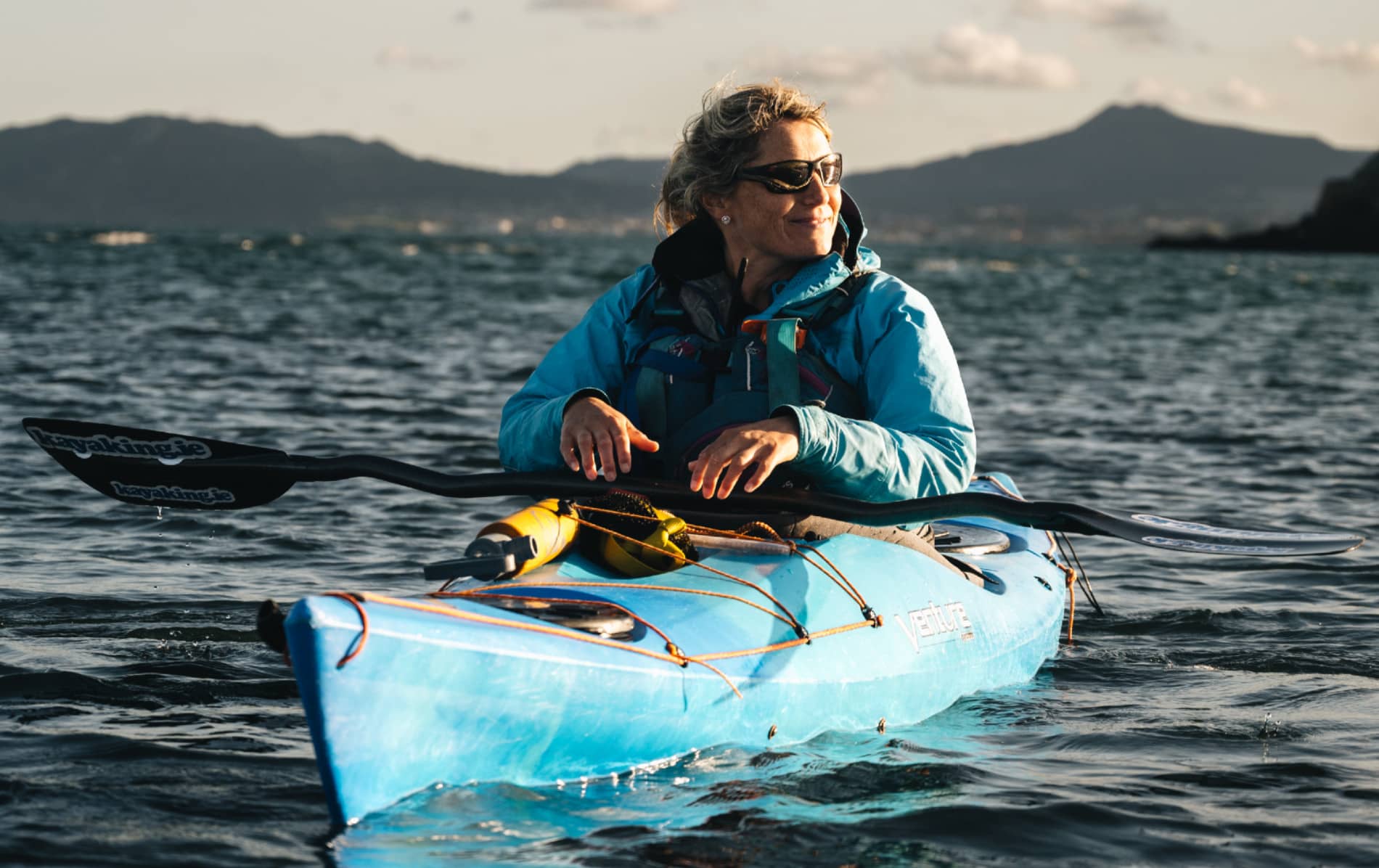 Kayaker wearing sunglasses sitting in blue kayak on Dublin Bay with Howth peninsula in the distance.