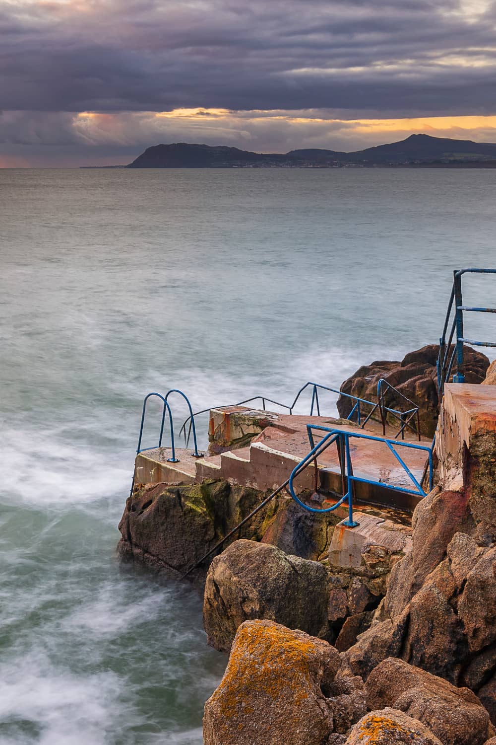 Concrete steps with blue railings descending into waves at the Vico Baths swimming spot, Killiney, Dublin.