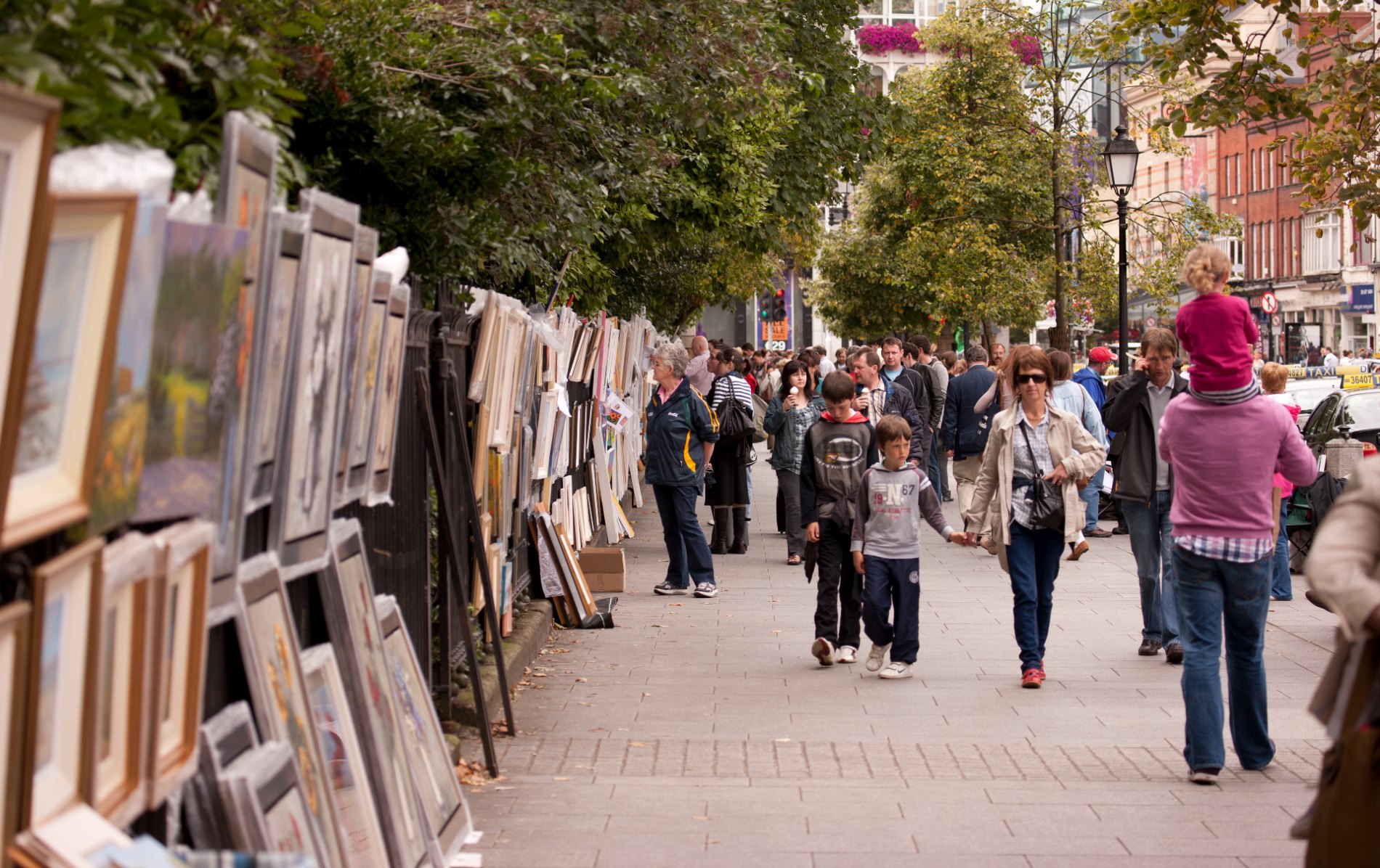 Crowds browsing outdoor art stalls with framed paintings along Dublin’s Merrion Square railings.