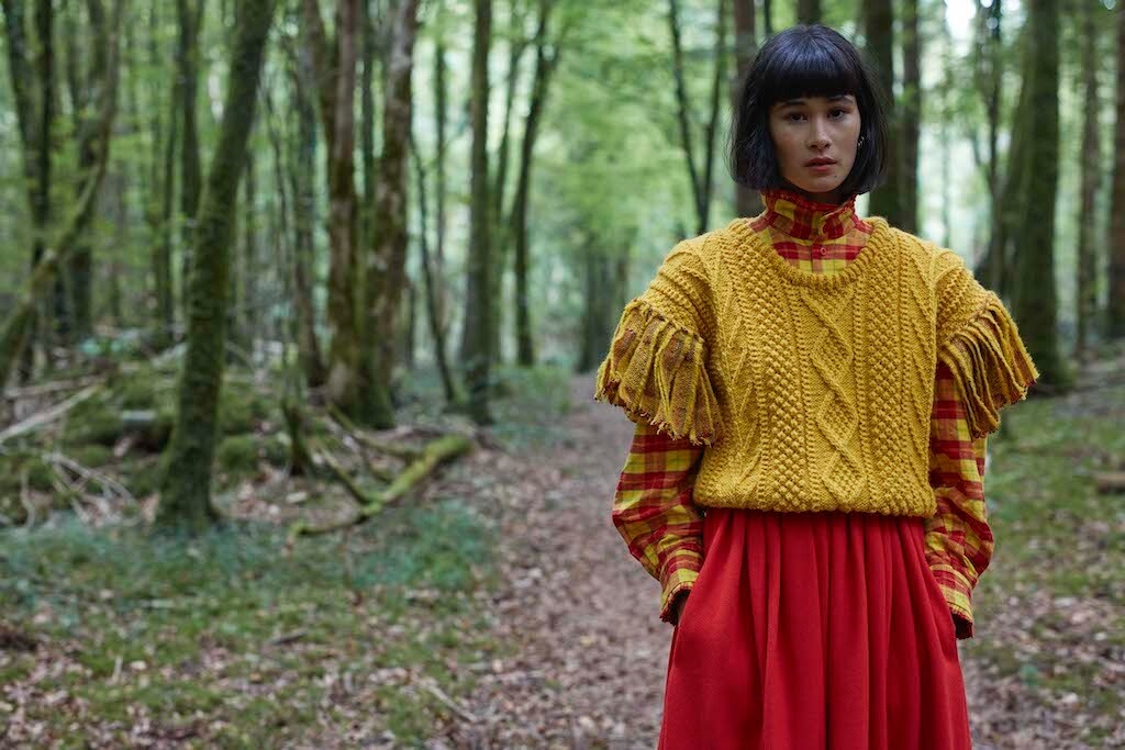 Model in forest path wearing a richly textured yellow wool sweater over a red plaid blouse and crimson skirt.
