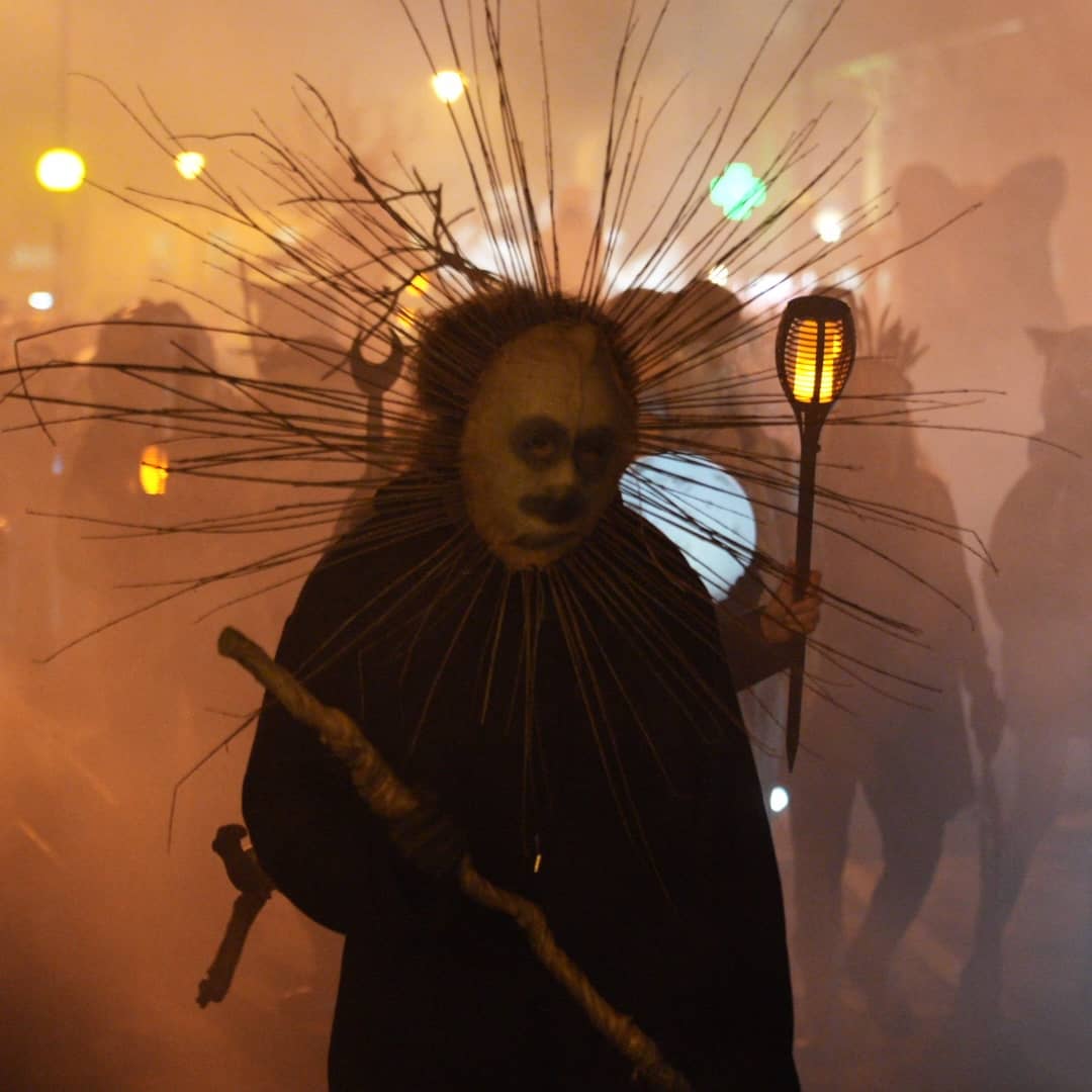 Costumed performer with spiked headdress and glowing lantern at an atmospheric festival procession.