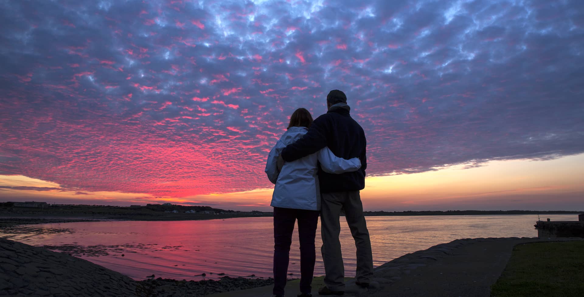 Couple standing arm in arm watching a vivid pink and purple sunset over calm waters off Burr Point, County Down.