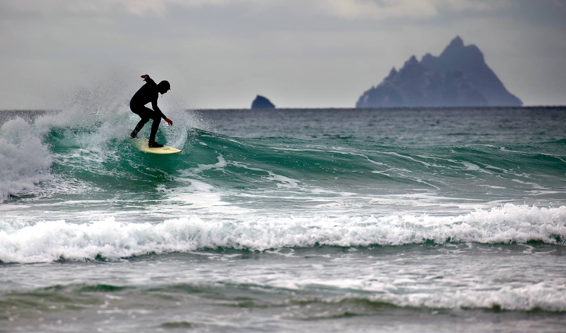 Surfer riding a wave off the Kerry coast with Skellig Michael island visible in the distance.