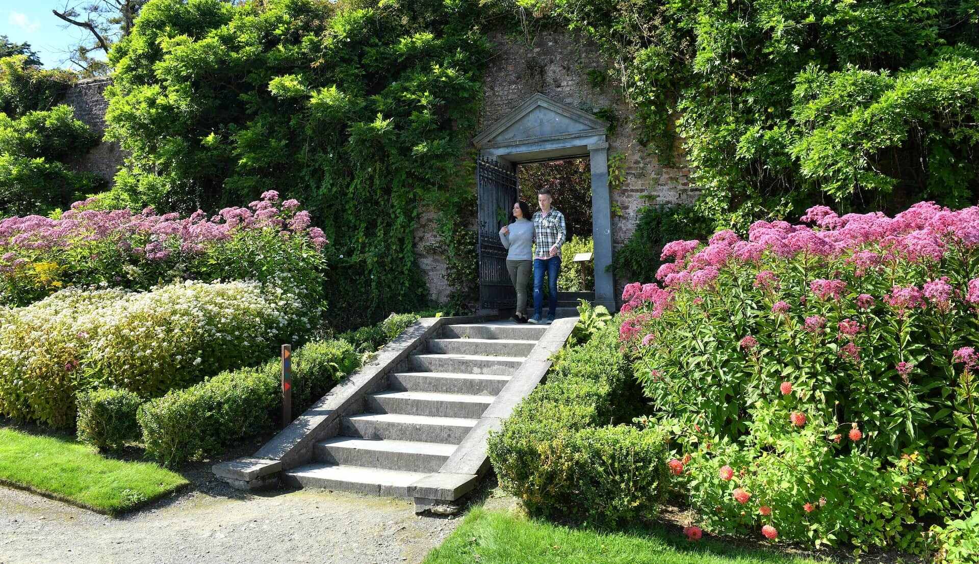 Couple walking through a stone archway surrounded by vibrant summer blooms at Mount Congreve Gardens, County Waterford.