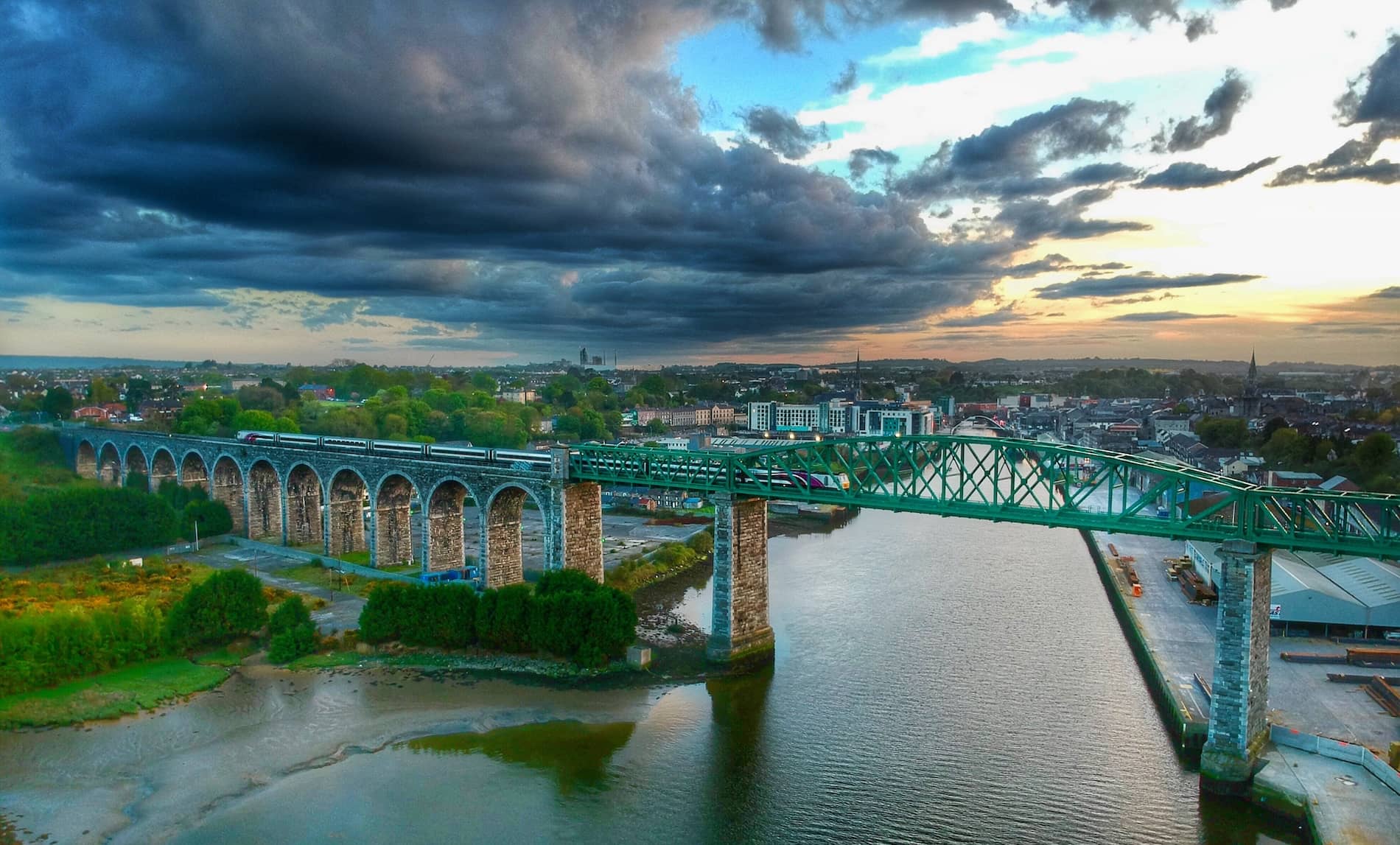 A train crosses the Boyne Viaduct at sunset, with dramatic clouds over the River Boyne and the town of Drogheda in the background.