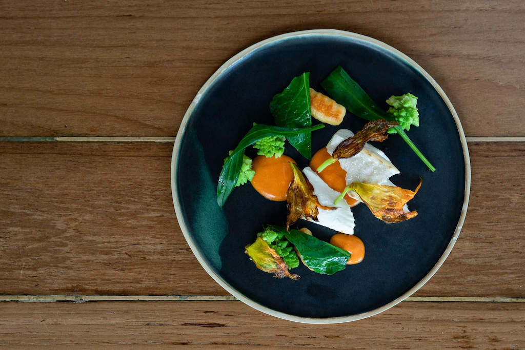 A modern dish of seasonal vegetables artfully arranged on a dark plate set on a wooden table.