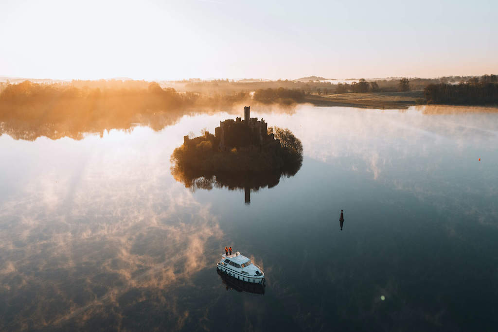 A castle ruin sits on a small wooded island surrounded by calm water and morning mist – a cruiser nearby..