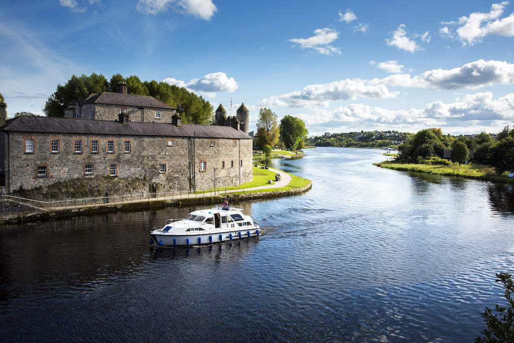 A cruiser boat passes a historic stone castle on a calm river beneath a blue sky.