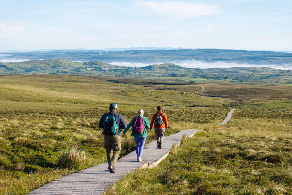 Walkers follow a wooden boardwalk across open bogland with hills and a lake in the distance.