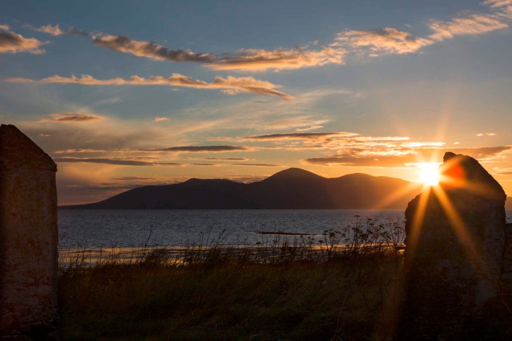 Sunset over the sea viewed between two stone ruins, with the Mourne Mountains in silhouette.