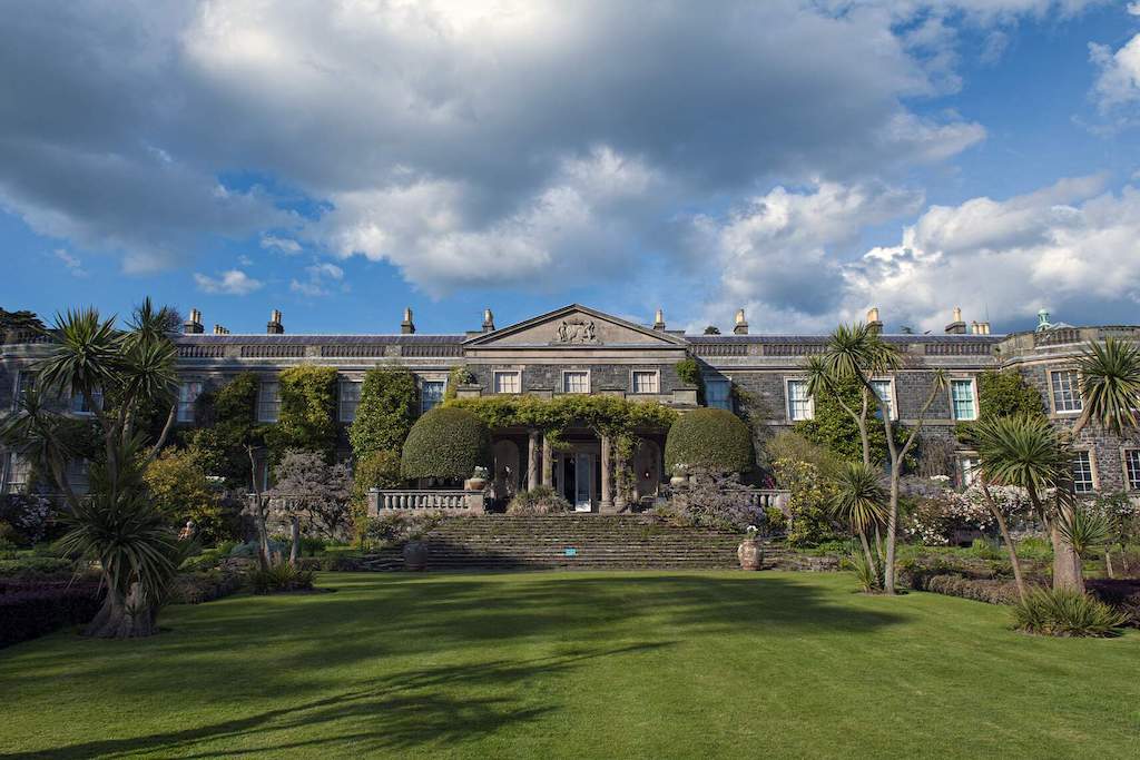 Elegant Georgian house with manicured gardens and palm trees under a blue sky with clouds.