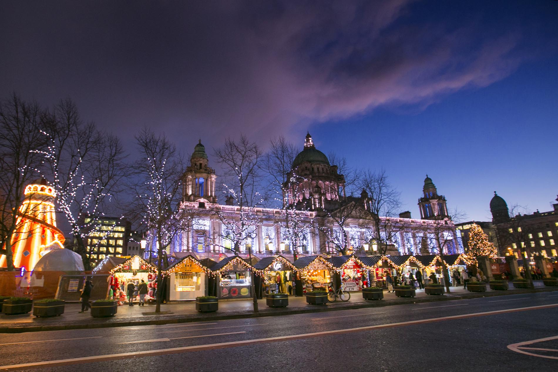 Belfast City Hall lit up at dusk with Christmas market stalls and festive lights in front.