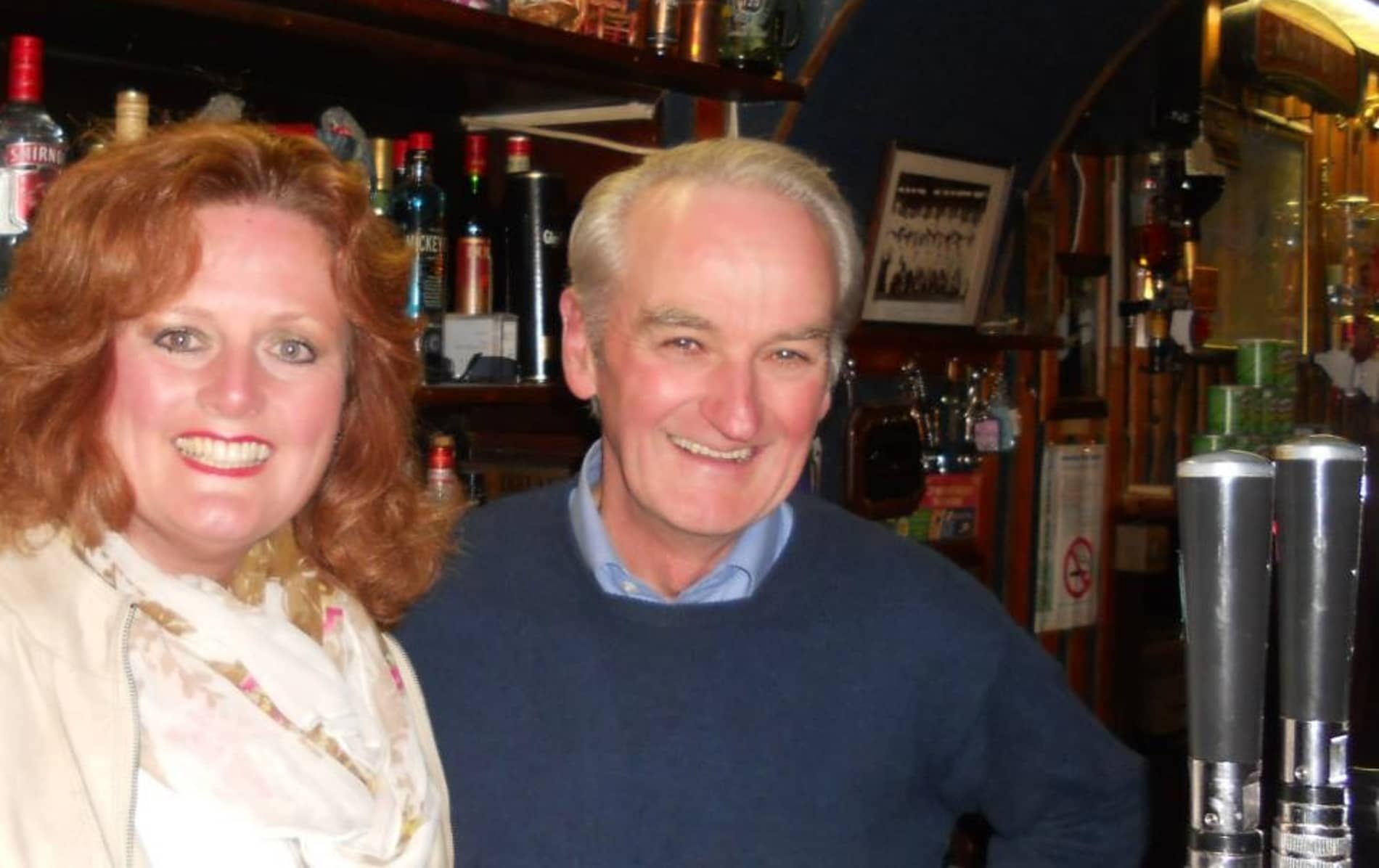 Two people smiling inside a traditional Irish pub with bottles and taps behind them.