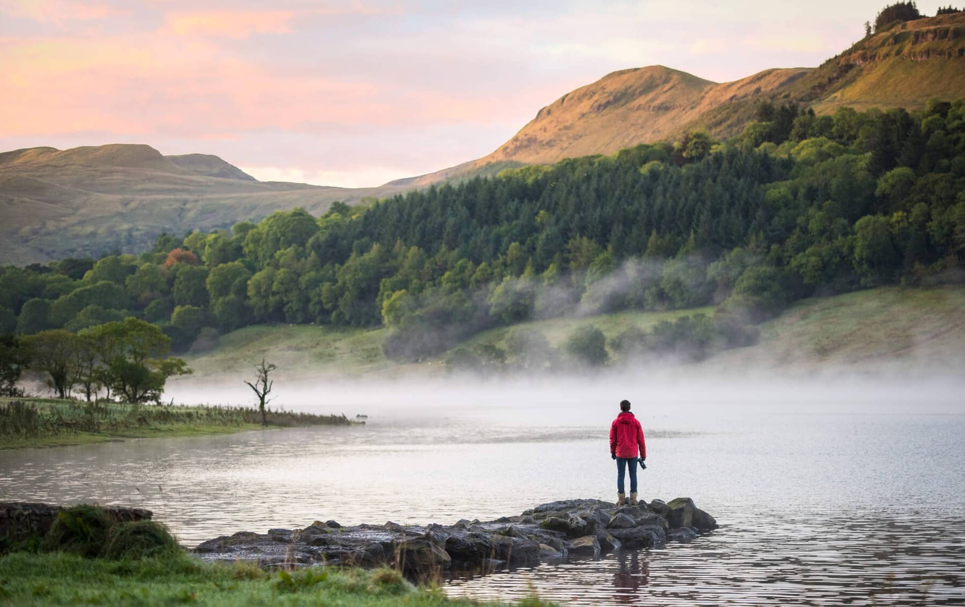 Lone figure in red jacket standing by misty lake with mountains in the background in Ireland.