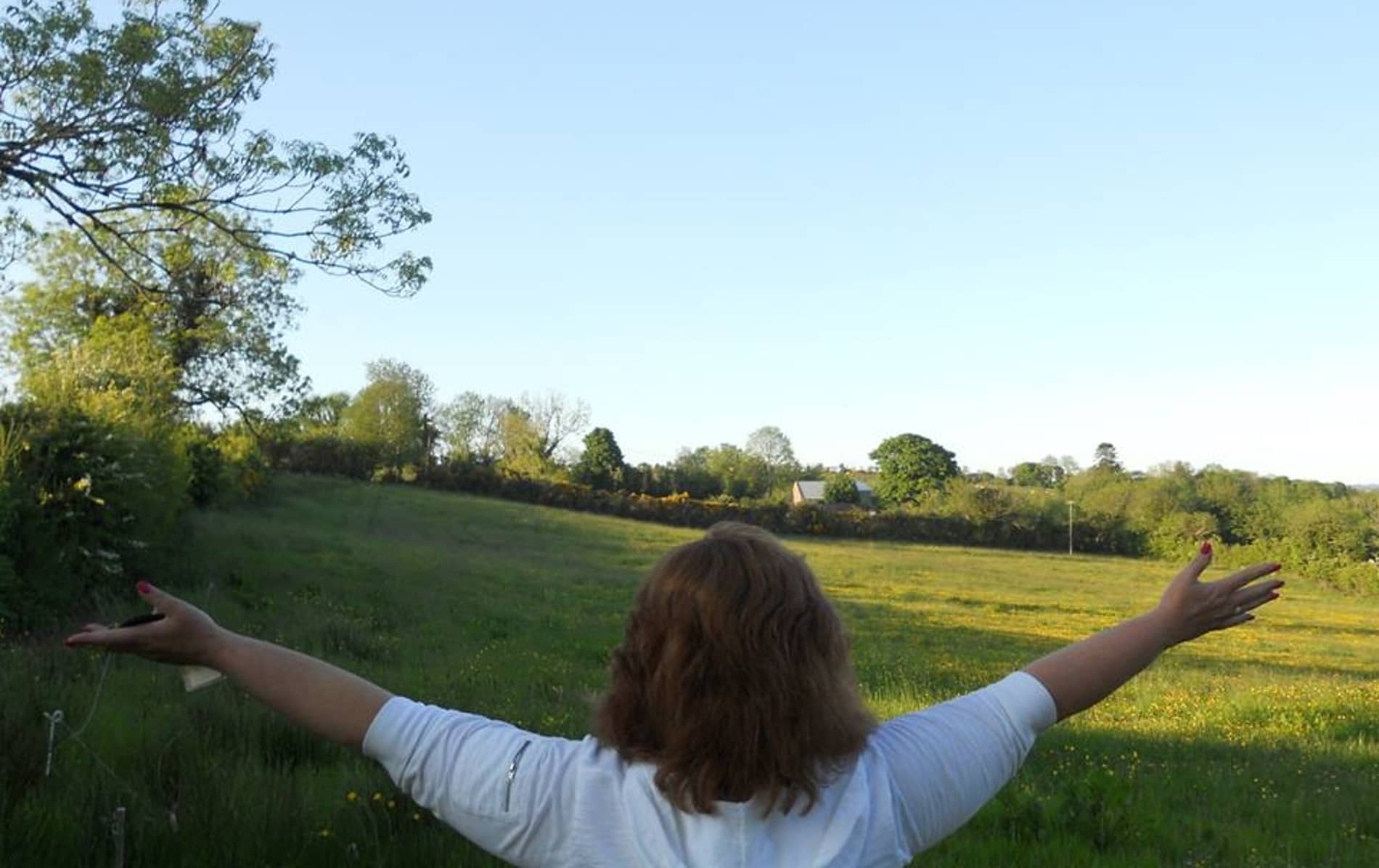 Woman with arms outstretched facing open green fields in Ireland.