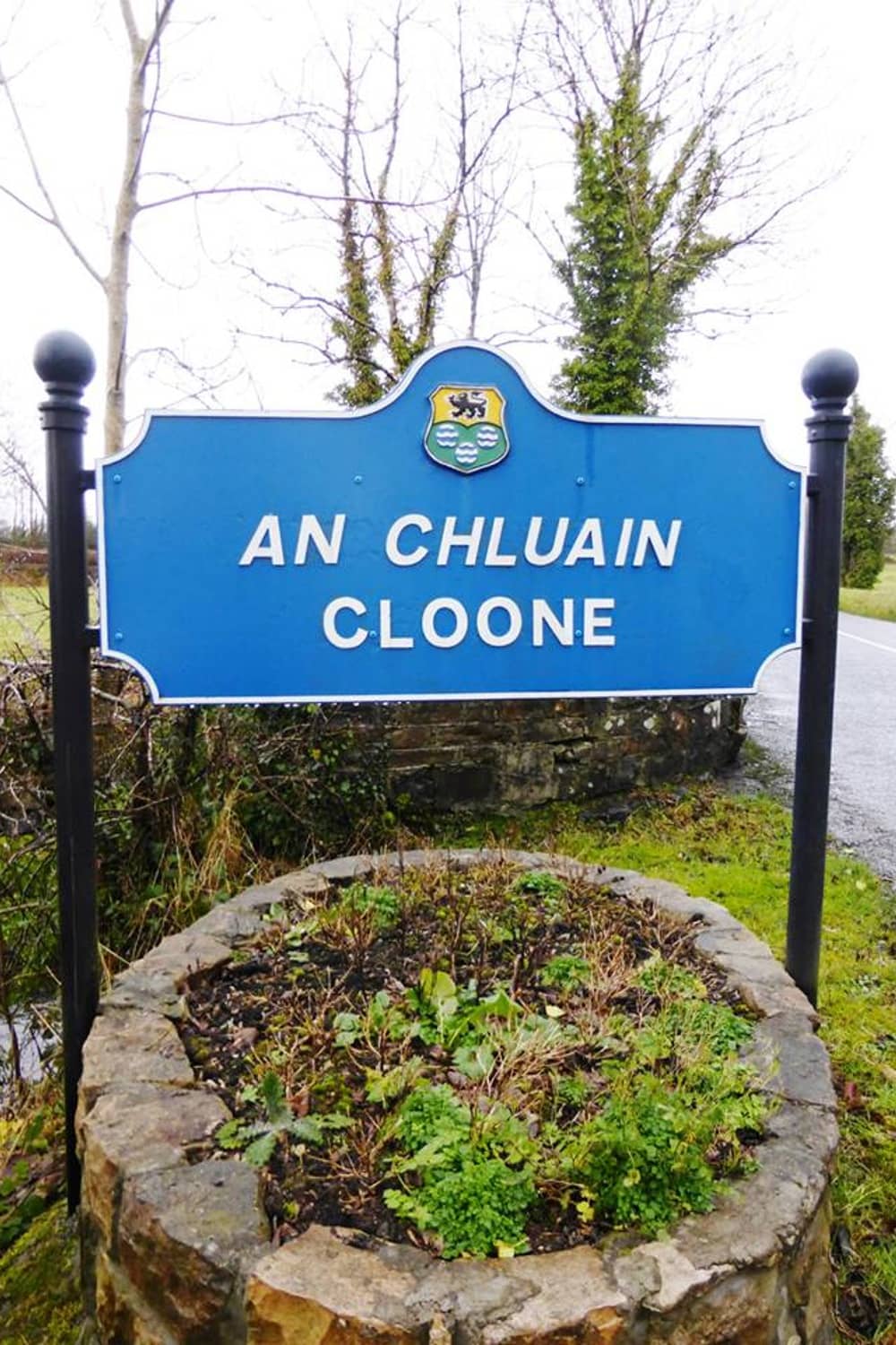 Roadside sign reading 'An Chluain Cloone' with county crest in Ireland.