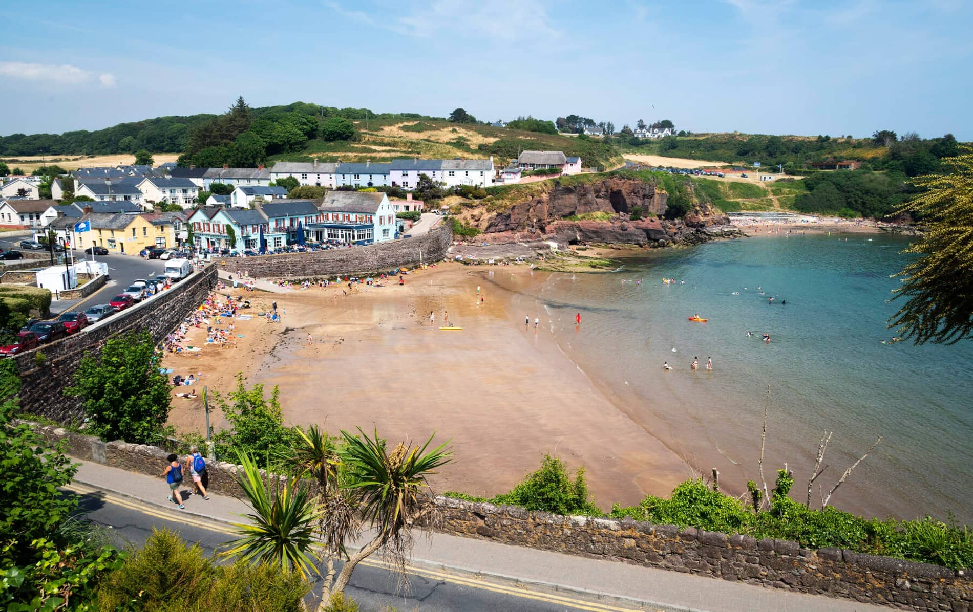 Dunmore East beach in County Waterford with people sunbathing and swimming near colourful buildings by the harbour.
