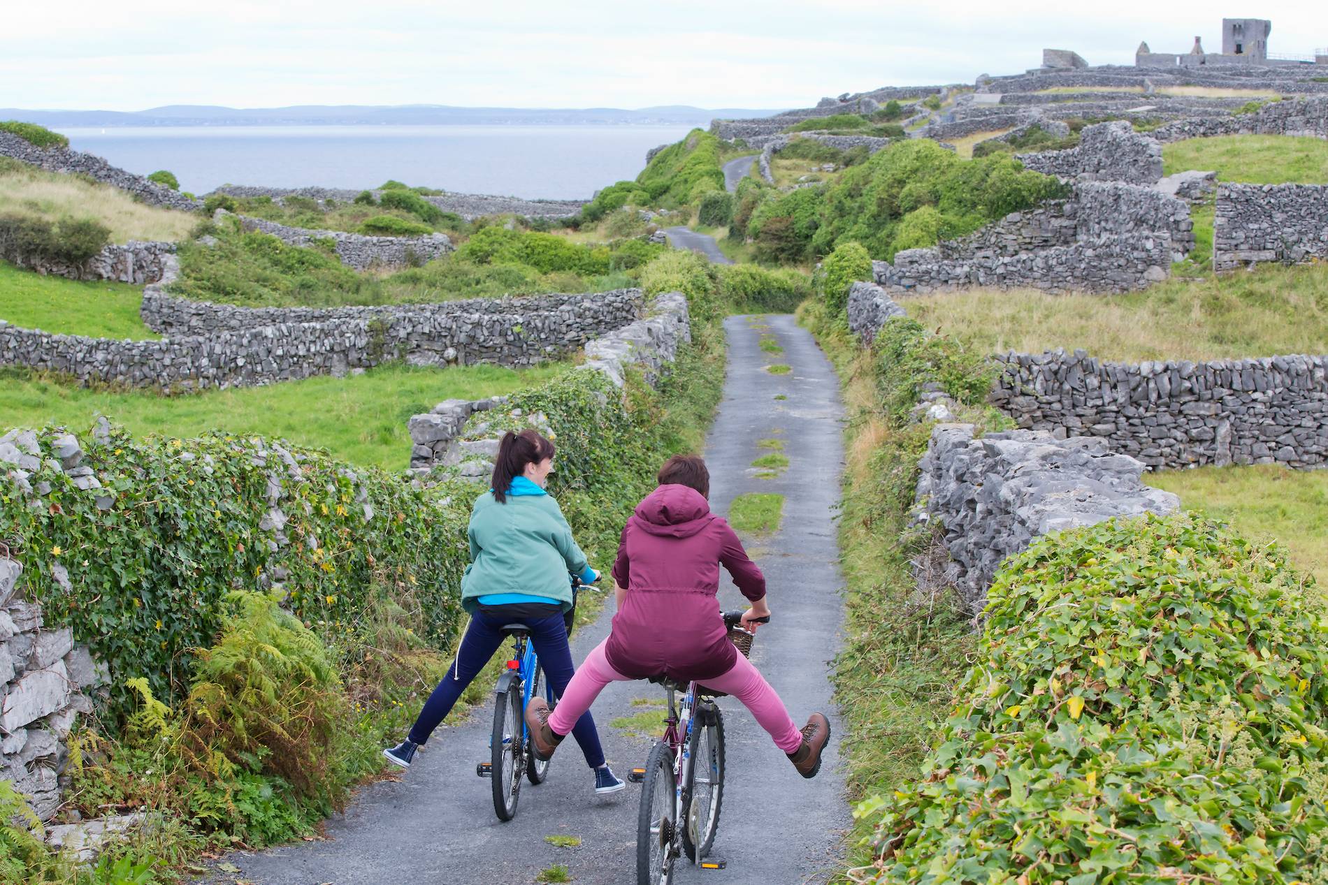 Two cyclists, one playfully with legs out, ride along a narrow path flanked by stone walls on the Aran Islands, Galway.