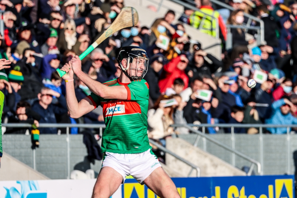 Hurling player in red and green kit swings hurley to strike sliotar during Cork senior final as crowd watches on.