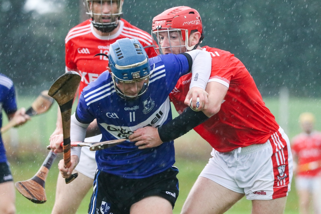 Player in blue jersey breaks past red-shirted defender during a wet hurling championship clash in Cork.