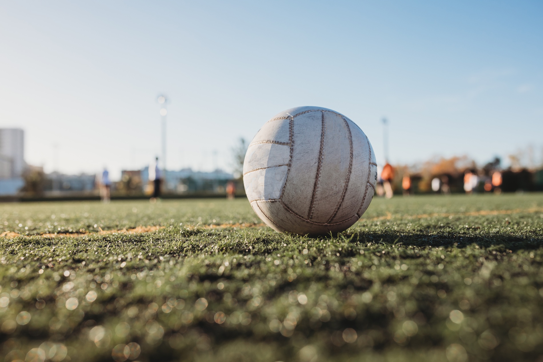 Gaelic football on sunlit pitch with blurred players training in the background at a GAA grounds.