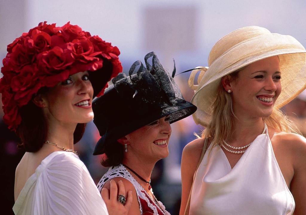 Three women in elegant hats and dresses smile while attending Ladies’ Day at the Galway Races.