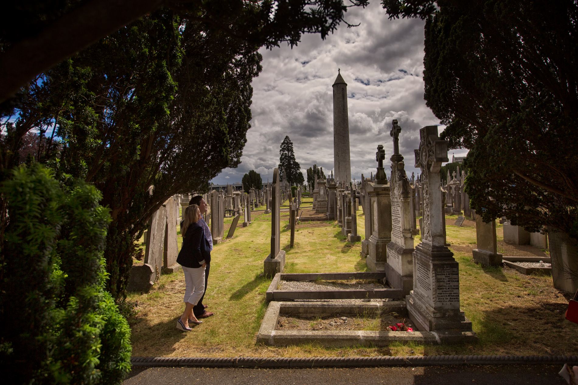 Visitors walking among historic gravestones at Glasnevin Cemetery, Dublin, with the tall round tower in the background.