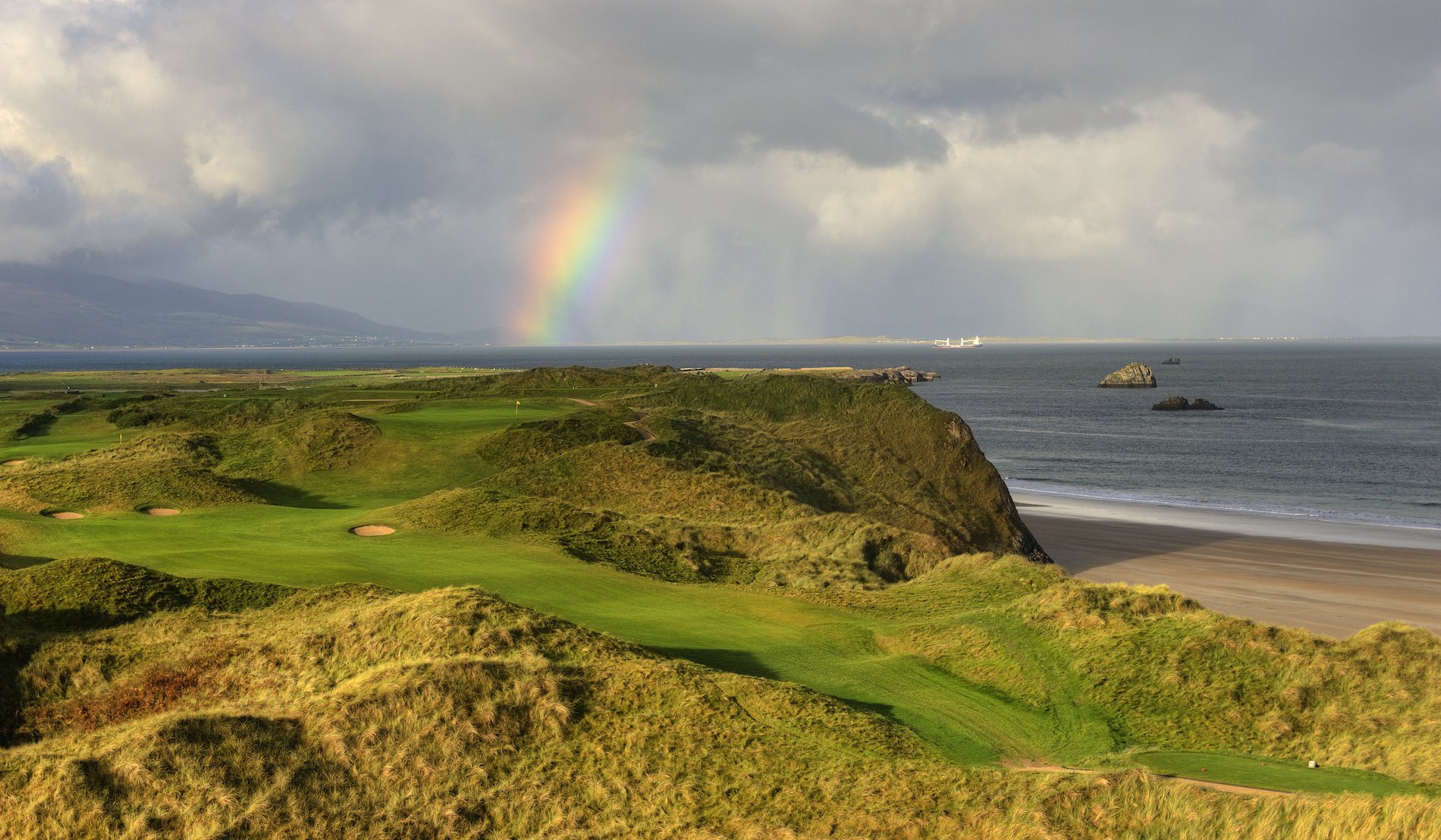Coastal golf course in Ireland with a rainbow over the sea and rolling fairways.