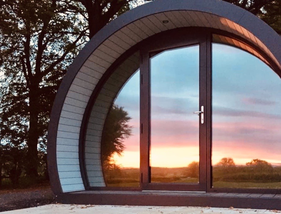 A curved glamping pod at Thornfield Farm, County Antrim, reflects the pink sunrise and rural landscape.