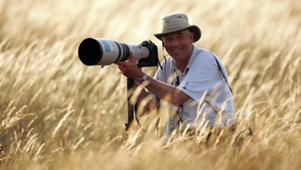 Golf photographer David Cannon kneels in tall grass with a long lens camera, smiling as he takes aim.