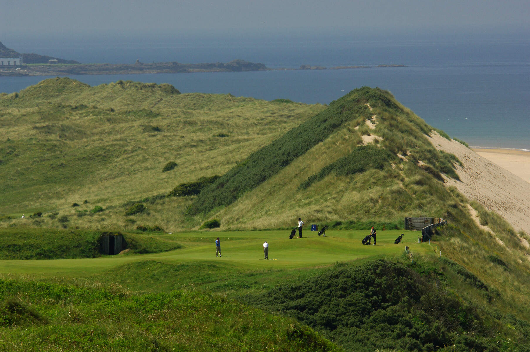 Sand dunes and fairways meet the Atlantic coast in a wide view of an Irish links course under blue skies.