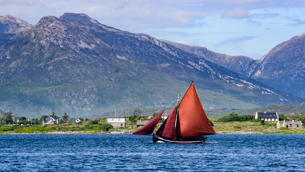 Traditional Galway Hooker boat with red sails sailing past cottages and Connemara mountains under blue sky.