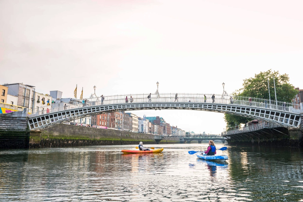 Two people kayaking the River Liffey under the Ha’penny Bridge with Dublin city buildings in the background.