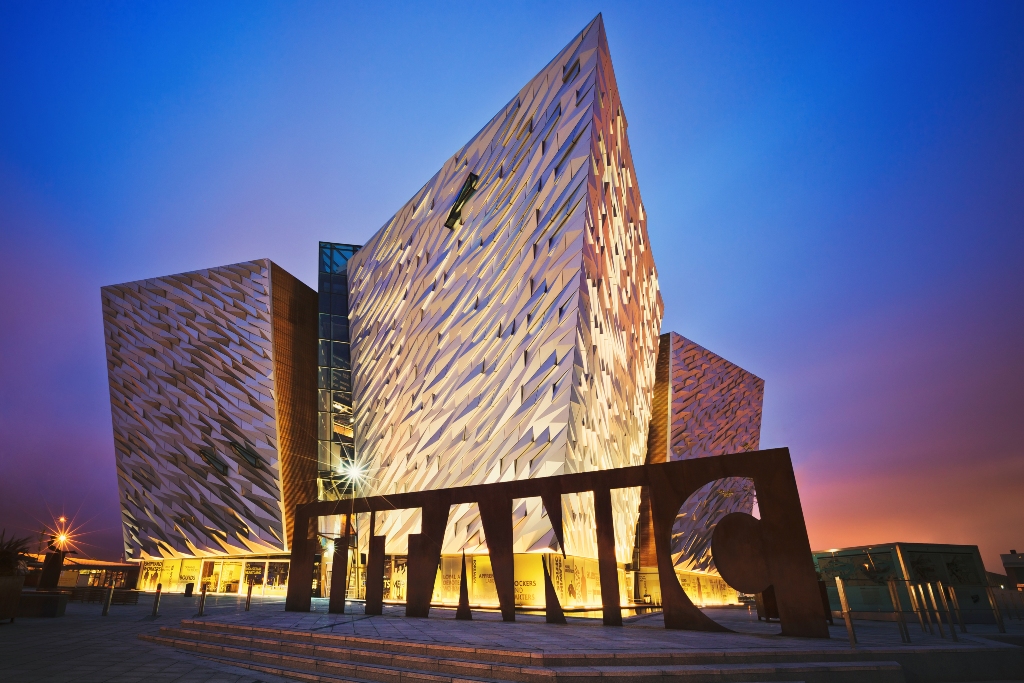 Titanic Belfast museum’s angular silver facade and large TITANIC sign illuminated at dusk in Belfast’s Titanic Quarter.
