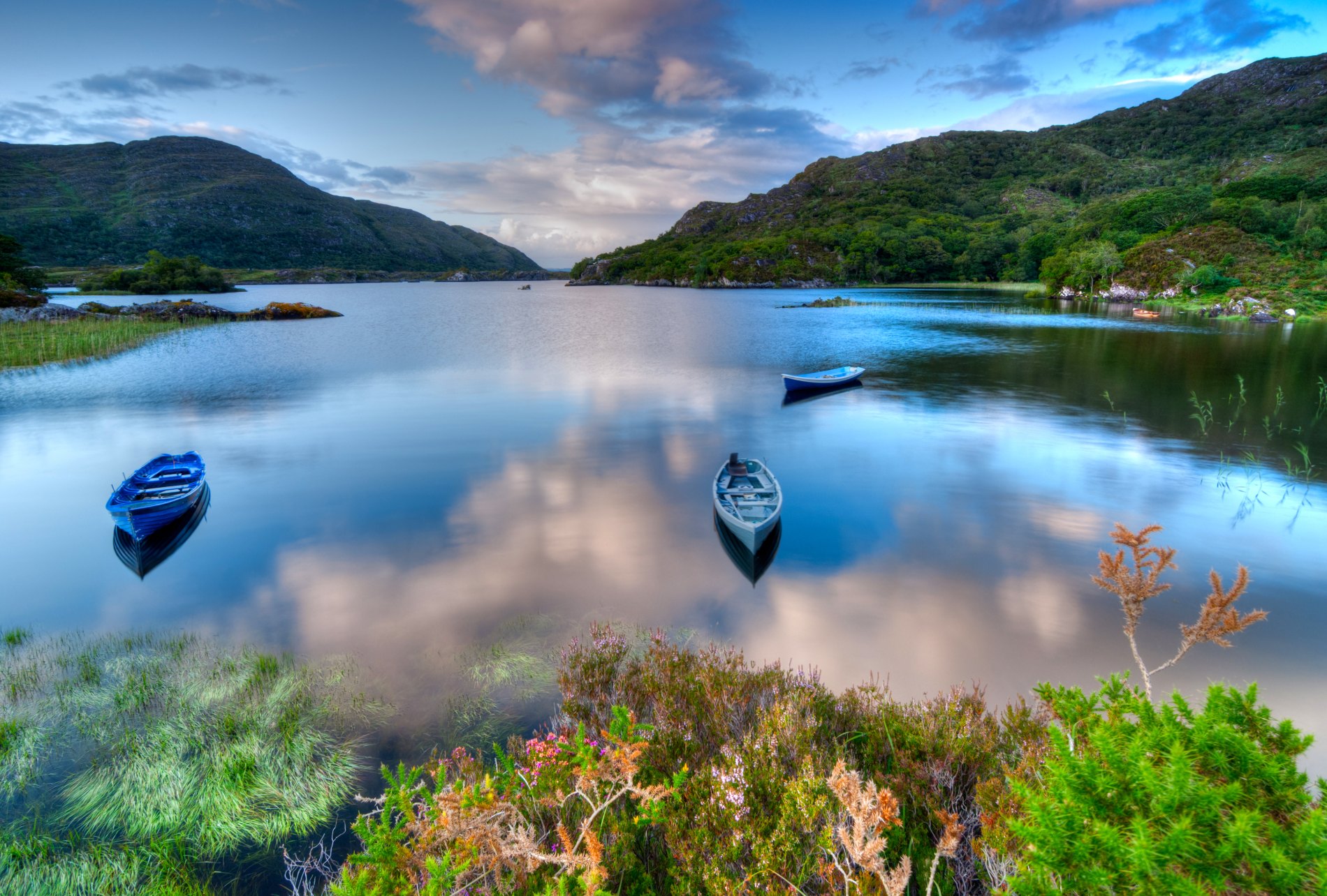 Calm lake in Killarney with wooded hills reflecting in water and small boats moored along grassy shore.