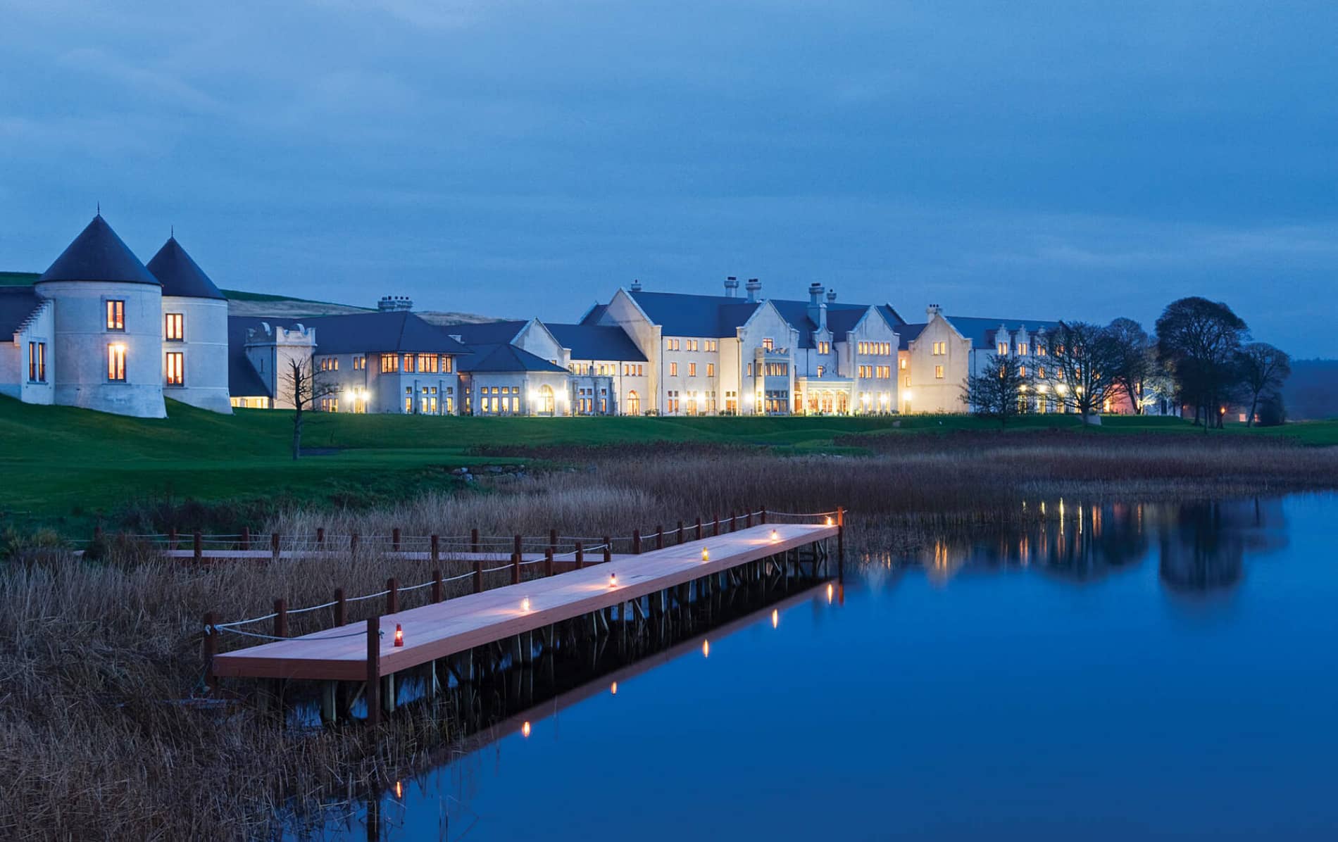 Lough Erne Resort at dusk in County Fermanagh, with warm lights glowing across its lakeside setting and private boardwalk.