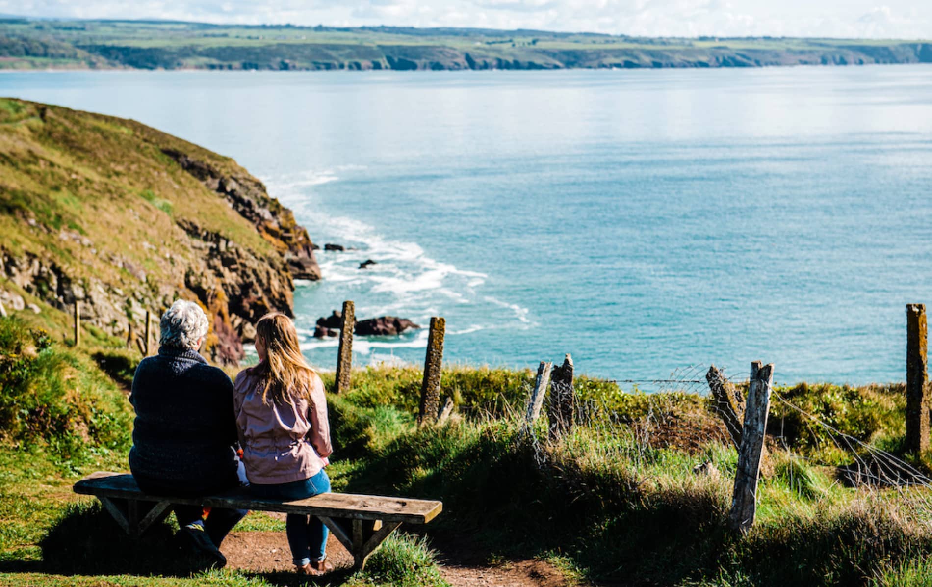Two people sitting on a clifftop bench overlooking Ardmore Bay, County Waterford, with waves breaking below.