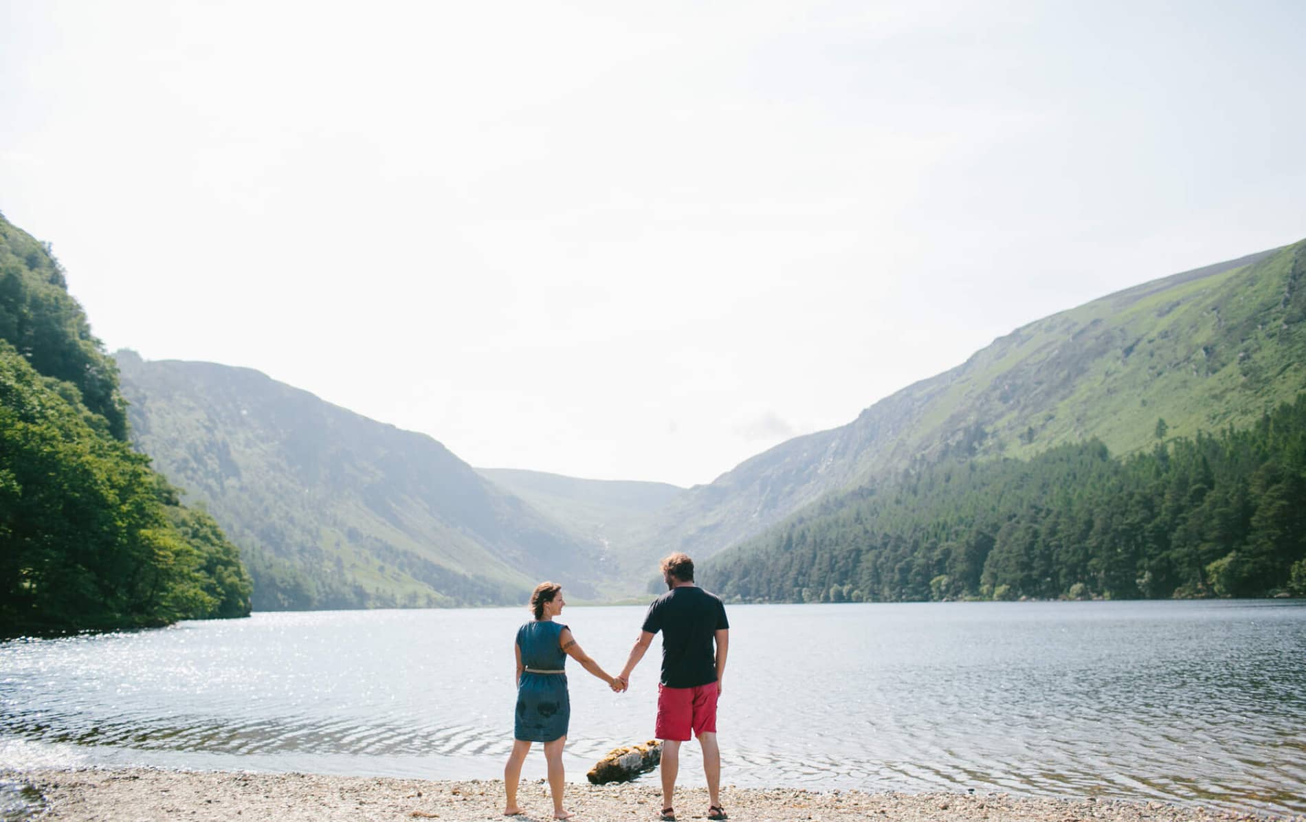 Couple holding hands by a lake at Glendalough, County Wicklow, framed by forested mountains under a pale sky.