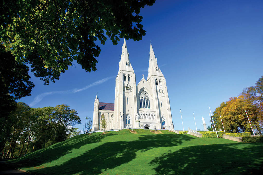 Twin-spired St Patrick’s Cathedral rising above a sunlit green hill and framed by trees in Armagh city.