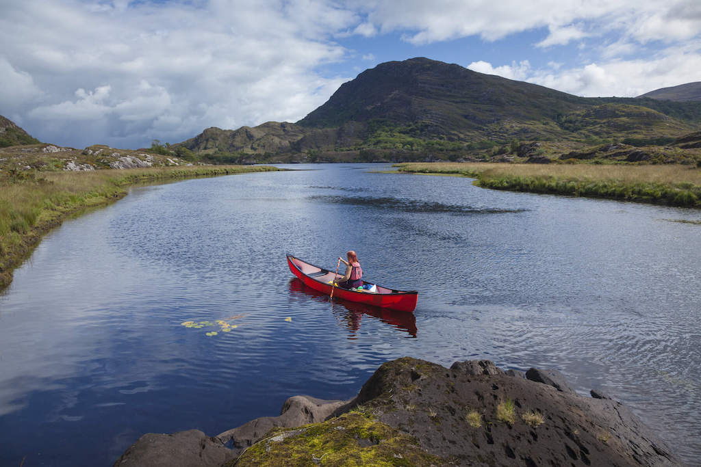Solo paddler in a red canoe on a tranquil lake surrounded by rugged mountains in Killarney National Park.