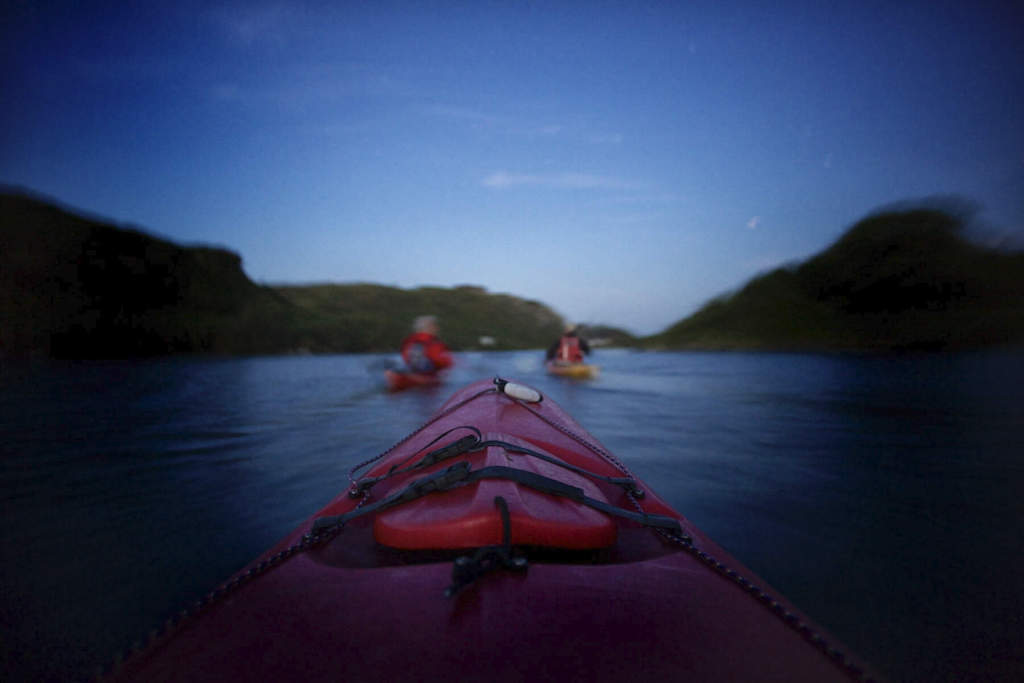 Kayakers paddling across the still waters of Lough Hyne, County Cork at twilight, surrounded by forested hills.