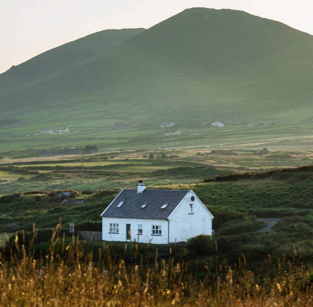 White cottage in the Ring of Kerry set against green fields and a softly lit mountain backdrop at sunset.