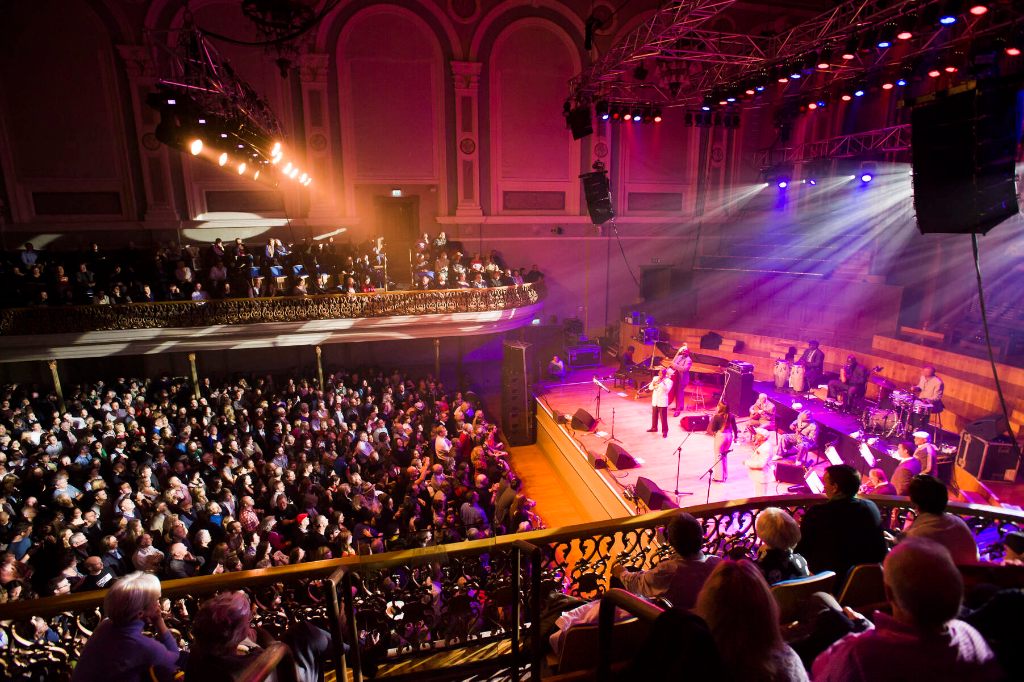 Packed Ulster Hall in Belfast with a band performing on stage under bright purple and yellow lights.
