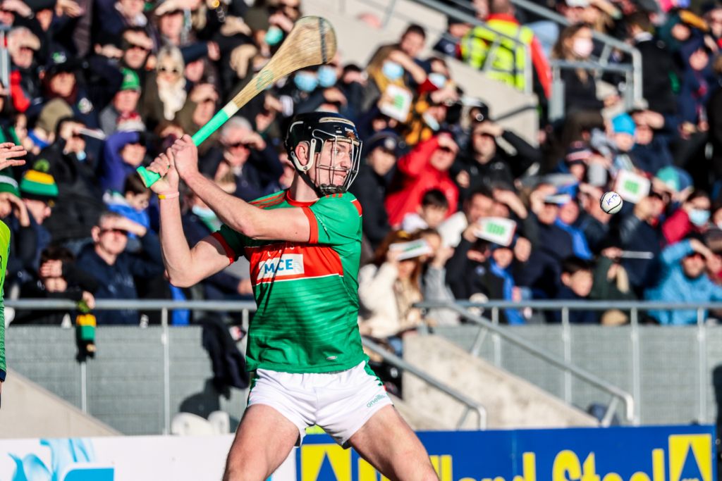 Hurler in green and red jersey striking a sliotar with a wooden stick during a match in Ireland.