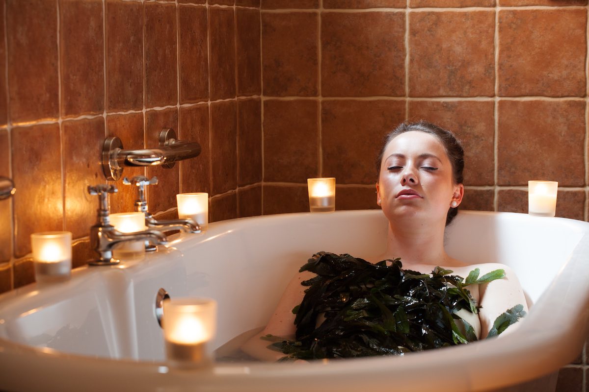 Person reclining in bath with seaweed on chest, eyes closed, surrounded by lit candles on ledge.
